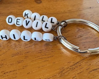 Peut inclure: Un porte-clés fait à la main avec des perles blanches épelant "KILL DEVIL HILLS" en lettres noires. Le porte-clés a un anneau en métal argenté et est présenté sur une surface en bois.