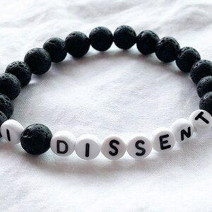 Peut inclure: Un bracelet en pierre de lave noire avec des perles blanches qui &eacute;pellent le mot "DISSENT".
