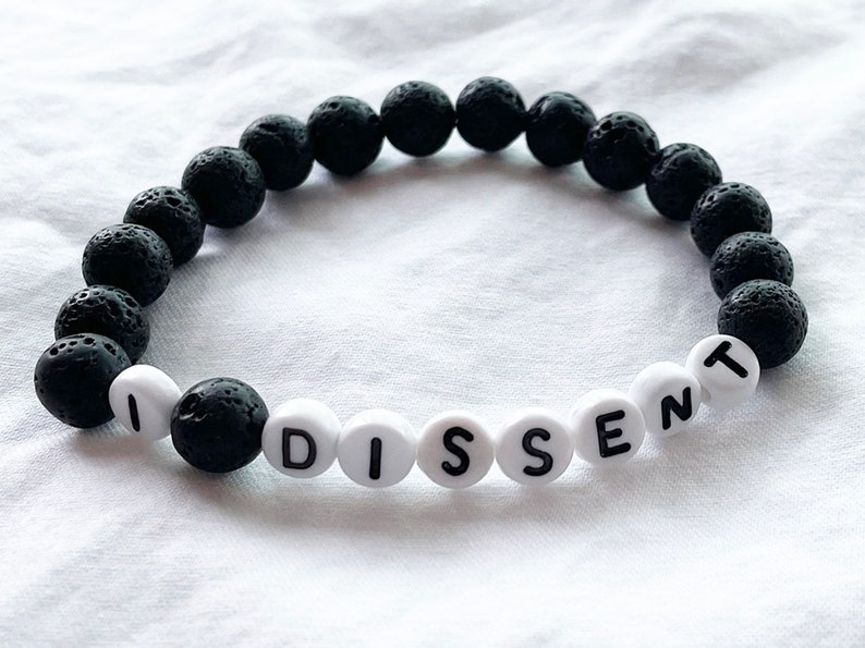 Peut inclure: Un bracelet en pierre de lave noire avec des perles blanches qui &eacute;pellent le mot "DISSENT".