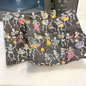 May include: Two black fabric pot holders with a skeleton band playing musical instruments pattern. The pot holders are folded and have a quilted texture.