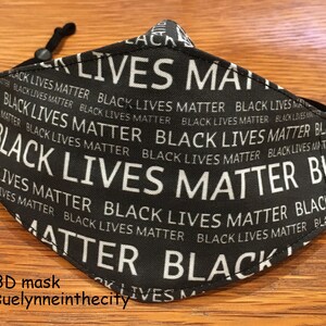 May include: Black face mask with the words "Black Lives Matter" repeated in white text on a black background. The mask has elastic ear loops.