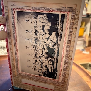 May include: Handmade journal with a vintage baseball theme. The cover features a black and white team photo, stitched with red thread. The journal pages are made from newspaper clippings, with a red and white rope spine and baseball charms.