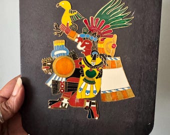 May include: A rectangular, dark-colored wooden plaque with a colorful, stylized depiction of a figure holding a yellow bird. The figure is adorned with a headdress and yellow, red, and green clothing. The artwork has gold accents.