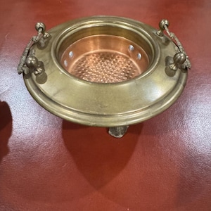 May include: A brass and copper serving dish with two handles. The dish has a round, shallow bowl with a copper insert. The dish is on a brown surface.