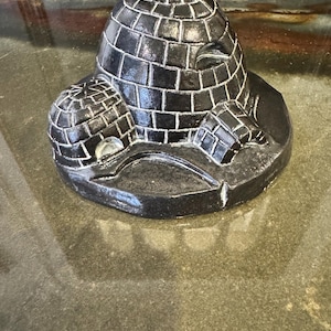 May include: A black ceramic igloo sculpture with a central dome and two smaller igloos on either side. The sculpture has a textured, brick-like pattern and is set on a dark surface. The top of the central dome has a starburst design.