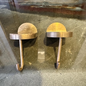 May include: Two antique brass hooks with a circular base and a curved hook. Each hook has a half-circle detail on top. The hooks are displayed on a dark, textured surface.