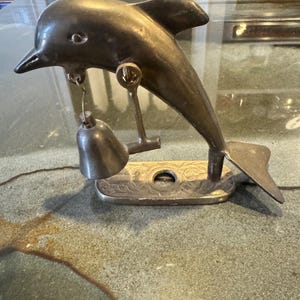 Könnte beinhalten: Eine bronzefarbene, delfinförmige Glocke mit einer kleinen Glocke, die an ihrem Maul hängt. Der Delfin ist auf einem rechteckigen Sockel mit einem verzierten Design montiert. Die Glocke ist an einer kleinen Kette aufgehängt.