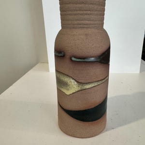 May include: A brown ceramic vase with a textured, ribbed neck and a cylindrical body. The body features horizontal bands of color, including black, yellow, and teal. The vase is a decorative item.