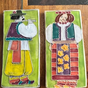 May include: Two rectangular ceramic tiles with folk art designs. One tile depicts a man playing a flute, wearing a hat, purple vest, and yellow trousers. The other tile shows a woman in a floral skirt and a white blouse. Both tiles have a green background.