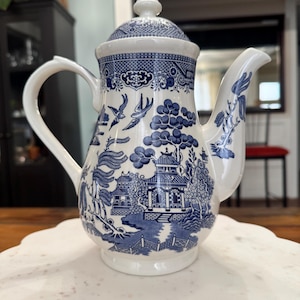 Può includere: Una teiera in ceramica bianca e blu con coperchio. La teiera presenta un classico motivo Willow con dettagliate illustrazioni blu di pagode, uccelli e paesaggi. Ha un manico curvo e un beccuccio per versare.