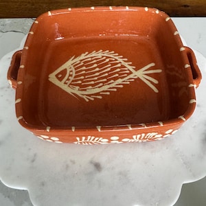 May include: A square terracotta baking dish with two side handles, featuring a white hand-painted fish motif at the center and white dash patterns along the rim. The exterior side displays additional white floral and leaf-like decorative accents.