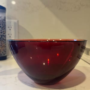 May include: A deep red glass bowl with a rounded shape. The bowl is empty and sitting on a white countertop.