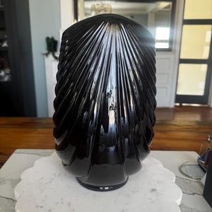 May include: A glossy black glass vase featuring a vertical ribbed or fluted pattern that radiates from the top centre down to the base. The vase has a rounded, shell-like silhouette with a smooth, reflective surface and a circular base.