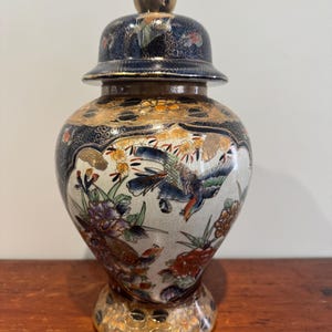 May include: An ornate ceramic jar with a lid, decorated with birds and floral patterns. The jar has gold detailing on the base and lid, with a dark blue and white body. The design is intricate and detailed, suitable for display.