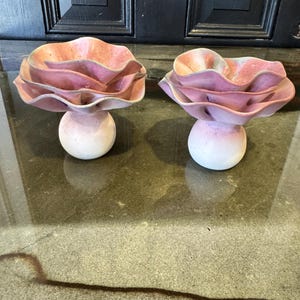 May include: Two ceramic flower sculptures. Each has layered pink and peach petals, and a white, spherical base. The background features a dark door and a reflective surface. The sculptures are decorative.