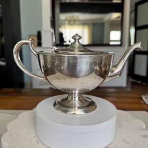 May include: A silver-toned metal teapot featuring a wide, rounded body, a curved handle, and a tapered spout. The teapot sits on a pedestal base and includes a matching lid with a small, pointed finial. The exterior surface is smooth and polished.