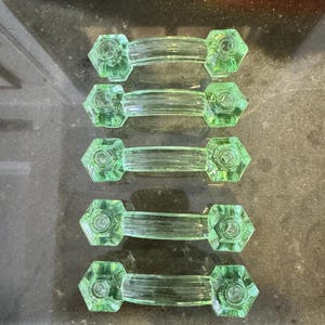May include: Five green glass cabinet pulls with a hexagonal design on each end. The pulls have a clear, ribbed center section. The glass has a slight green tint and a vintage aesthetic. The pulls are arranged in a row.