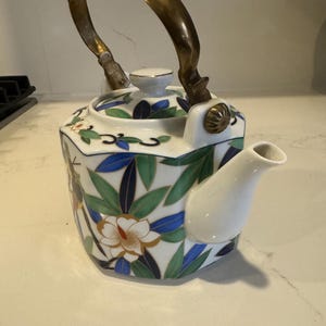 May include: A white porcelain teapot with a gold handle and floral design. The teapot has a geometric shape and features blue, green, and gold accents.