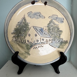 Peut inclure: Une assiette décorative ronde en céramique présentant une scène peinte à la main d'une maison nichée entre deux conifères sous deux nuages. L'assiette a un centre crème, un bord gris et le mot "Desiree" écrit en lettres cursives.