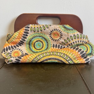 May include: A colorful clutch with a wooden handle. The fabric features a pattern of concentric circles in shades of orange, yellow, green, and blue, with black accents. The handle is a dark brown, curved shape.
