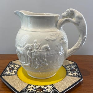 May include: A white ceramic pitcher featuring a relief-molded hunting scene with figures on horseback and a handle shaped like a dog. The pitcher rests on a hexagonal plate with a yellow center and a decorative border of black, white, and floral patterns.