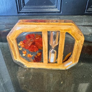 May include: A decorative, rectangular resin paperweight with a wooden frame. The paperweight contains a red flower, orange sprigs, and a small hourglass. The hourglass is silver and white. The frame is light brown.