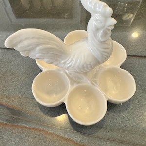 May include: A white ceramic egg holder in the shape of a flower with six egg cups. A detailed rooster figurine sits atop the holder. The egg holder is a decorative kitchen item.