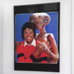 May include: A framed poster featuring Michael Jackson and E.T. The Extra-Terrestrial. Michael Jackson is wearing a red sweater with black stripes. E.T. is a light brown color with large blue eyes. The poster is in a black frame.