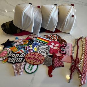 May include: An assortment of accessories including baseball caps with white tops and various colored brims, decorative patches with text and designs, and beaded and chain necklaces. The patches feature phrases like "Hot Girls", "Wine Time", and "Just Beach".
