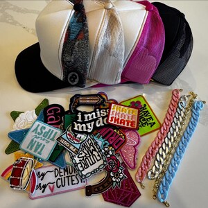 Peut inclure: Plusieurs casquettes de camionneur de couleurs variées, dont blanc, noir et rose, avec des dos en filet et des visières à motifs. Également, une pile d'écussons brodés avec du texte et des images, et plusieurs bracelets à maillons roses, argentés, dorés et bleus.