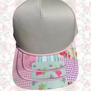 May include: A baseball cap featuring a solid light grey crown and a curved brim decorated with a multi-coloured patchwork pattern. The pattern includes pink and green floral motifs, light green stripes, and a pink and white checkered design.