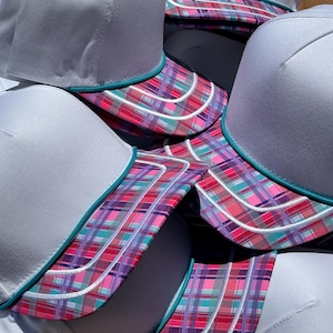 May include: A collection of white baseball caps featuring a vibrant plaid pattern on the brim in shades of pink, purple, and teal. Each cap includes a thin teal piping detail separating the white crown from the multicolored plaid brim.