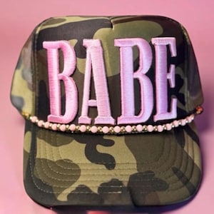Puede incluir: Gorra de camionero de camuflaje con la palabra "BABE" bordada en rosa. El sombrero presenta un ribete de cuentas a lo largo del borde y un diseño clásico de gorra de camionero.