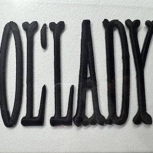 May include: Black embroidered text spelling out "OLD LADY" on a white background. The letters are thick and stylized, with decorative elements at the ends of some letters, giving a vintage or handcrafted feel.