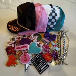 May include: A collection of colorful trucker hats with mesh backs, alongside decorative patches and chain necklaces. The hats feature various designs, including camouflage, pink, and checkered patterns. Patches include hearts, bows, and text.