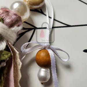 Puede incluir: Primer plano de un adorno decorativo con una cinta blanca y una cuenta de madera. El adorno tambi&eacute;n presenta una perla grande y una cuenta m&aacute;s peque&ntilde;a de color dorado. El fondo es una superficie blanca con l&iacute;neas negras.