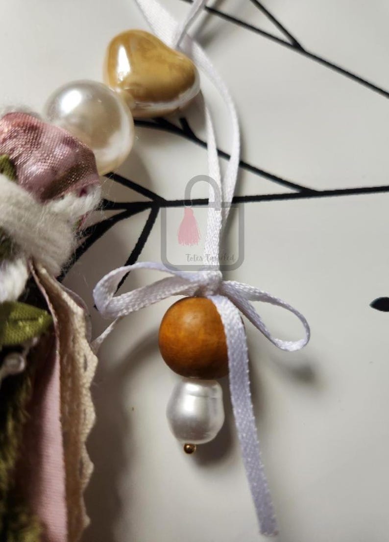 Puede incluir: Primer plano de un adorno decorativo con una cinta blanca y una cuenta de madera. El adorno tambi&eacute;n presenta una perla grande y una cuenta m&aacute;s peque&ntilde;a de color dorado. El fondo es una superficie blanca con l&iacute;neas negras.