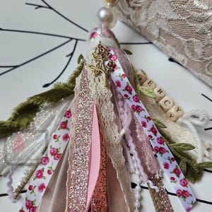 Puede incluir: Una borla decorativa con varias cintas, encajes y cuentas. La borla presenta cintas con estampados florales, purpurina y colores lisos en rosa, blanco y beige. Un dije dorado y cuentas de letras de madera deletrean "ERCY".