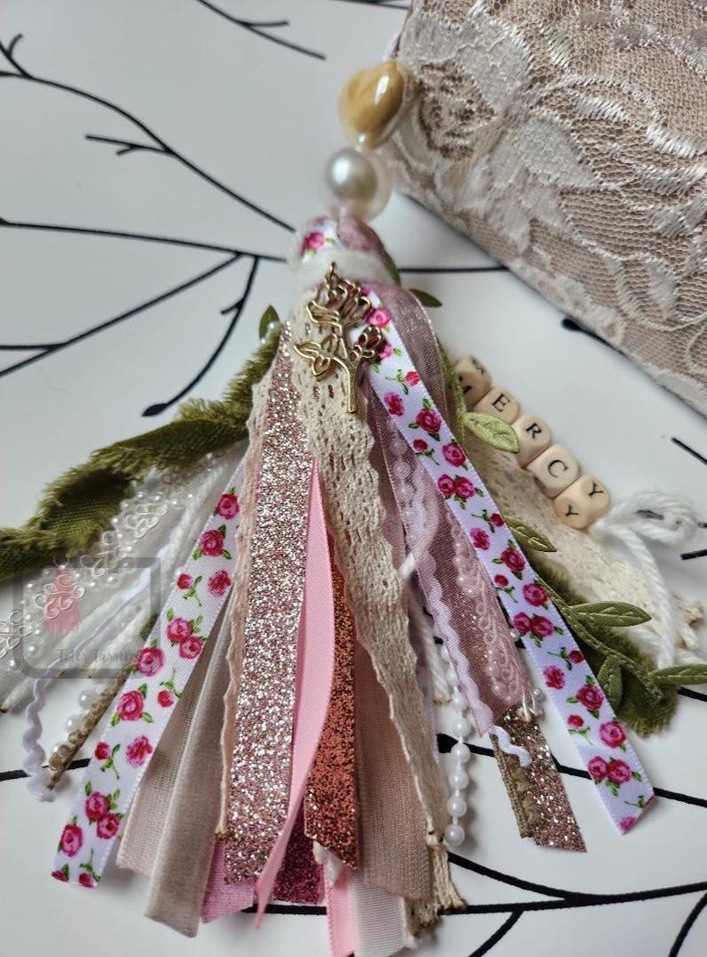 Puede incluir: Una borla decorativa con varias cintas, encajes y cuentas. La borla presenta cintas con estampados florales, purpurina y colores lisos en rosa, blanco y beige. Un dije dorado y cuentas de letras de madera deletrean "ERCY".