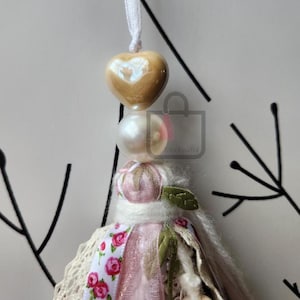 Puede incluir: Un adorno de borla hecho a mano con una cuenta en forma de coraz&oacute;n y una perla. La borla presenta una cinta floral rosa, encaje y una peque&ntilde;a hoja verde. El adorno est&aacute; suspendido por una cinta blanca.