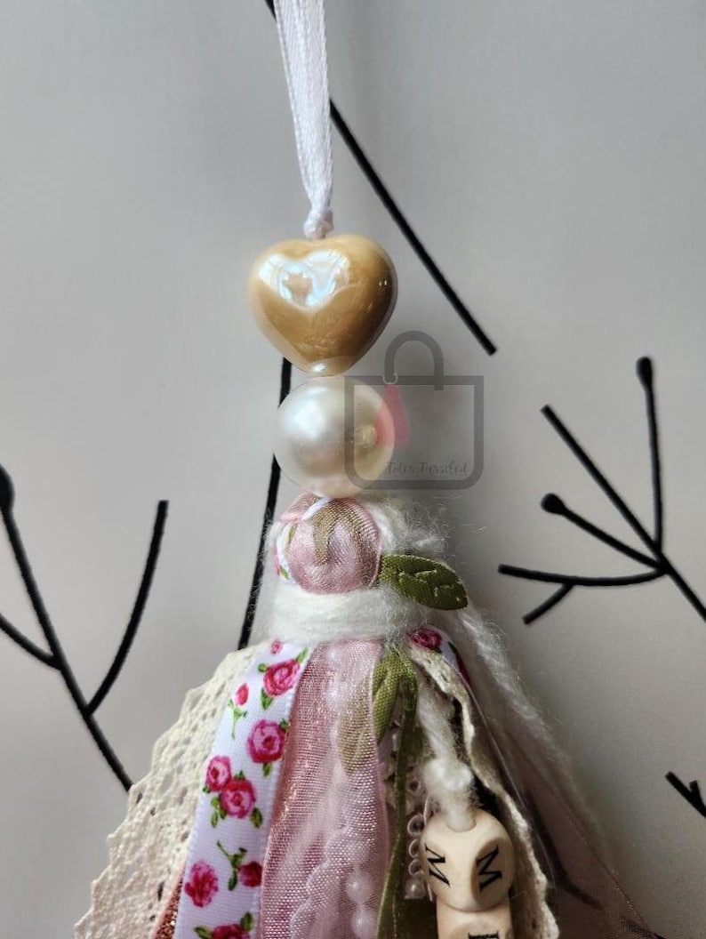 Puede incluir: Un adorno de borla hecho a mano con una cuenta en forma de coraz&oacute;n y una perla. La borla presenta una cinta floral rosa, encaje y una peque&ntilde;a hoja verde. El adorno est&aacute; suspendido por una cinta blanca.