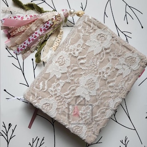 Puede incluir: Un diario beige con una superposici&oacute;n de encaje floral blanco. Una borla decorativa con cintas rosas, doradas y blancas, encaje y cuentas cuelga del diario. La palabra "MERCY" est&aacute; escrita en peque&ntilde;as cuentas de madera.