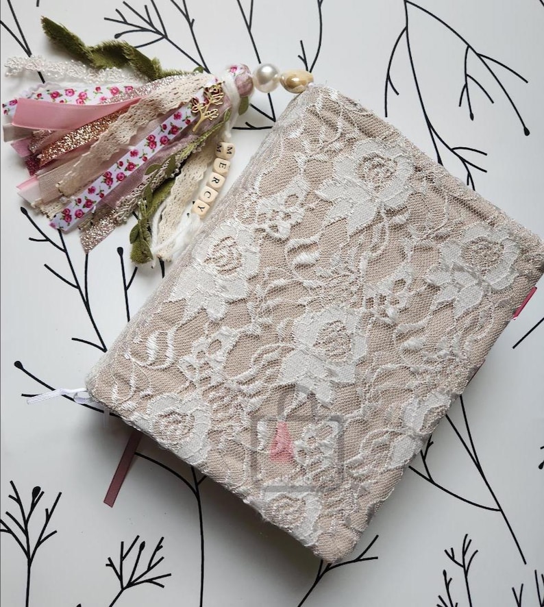 Puede incluir: Un diario beige con una superposici&oacute;n de encaje floral blanco. Una borla decorativa con cintas rosas, doradas y blancas, encaje y cuentas cuelga del diario. La palabra "MERCY" est&aacute; escrita en peque&ntilde;as cuentas de madera.