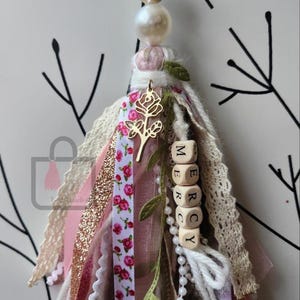 Puede incluir: Una borla artesanal con una cuenta dorada en forma de coraz&oacute;n, perla y detalles florales. La borla presenta cintas rosas, blancas y doradas, un dije de rosa dorada y cuentas de letras de madera que deletrean "MERCY". Art&iacute;culo decorativo.