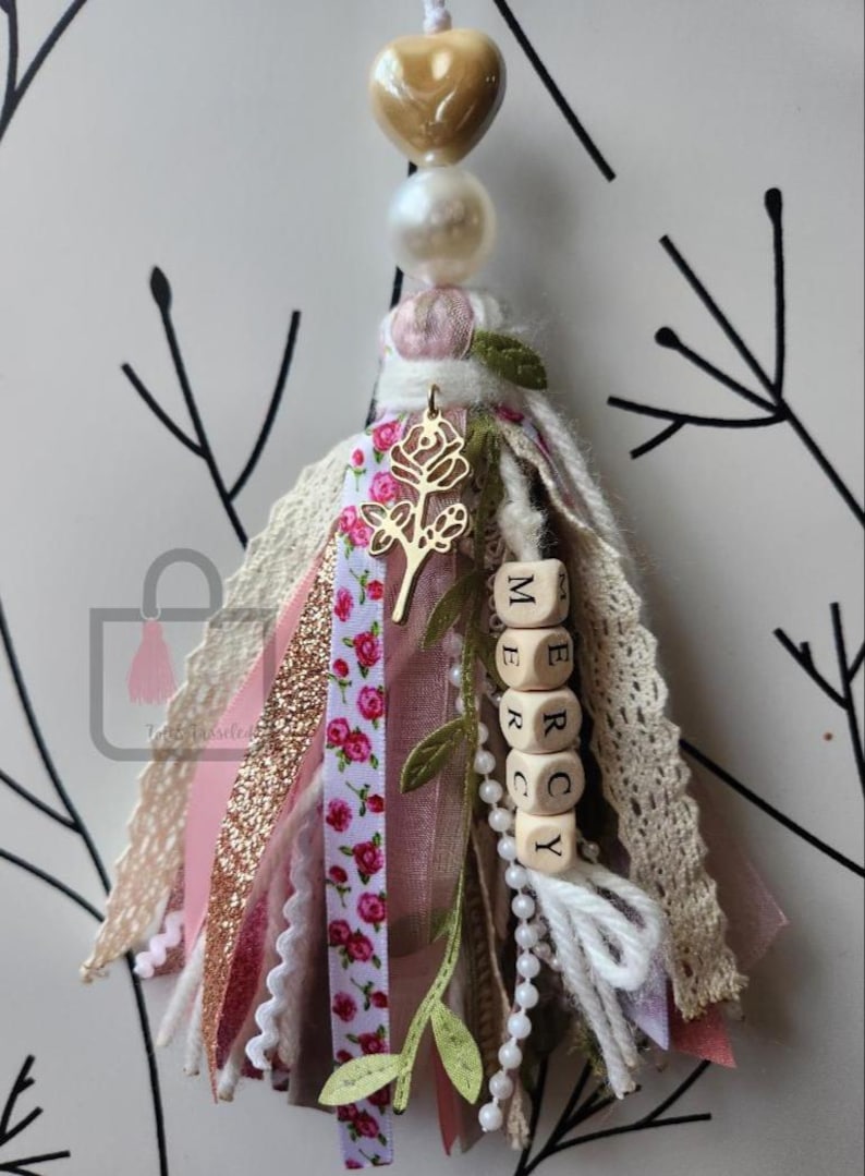 Puede incluir: Una borla artesanal con una cuenta dorada en forma de coraz&oacute;n, perla y detalles florales. La borla presenta cintas rosas, blancas y doradas, un dije de rosa dorada y cuentas de letras de madera que deletrean "MERCY". Art&iacute;culo decorativo.