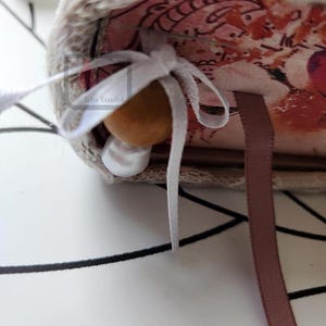 Puede incluir: Primer plano de un diario hecho a mano con un interior estampado floral. El diario est&aacute; atado con una cinta blanca y presenta una cuenta de madera y una perla. Una cinta de marcador marr&oacute;n es visible.