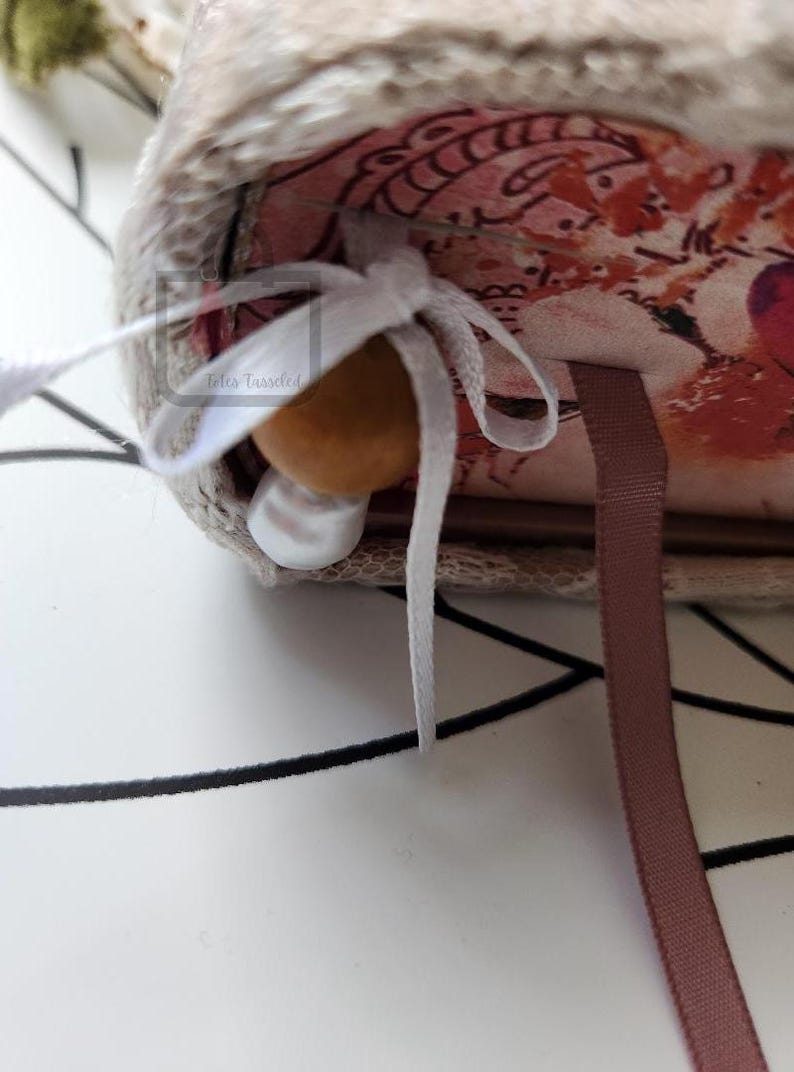 Puede incluir: Primer plano de un diario hecho a mano con un interior estampado floral. El diario est&aacute; atado con una cinta blanca y presenta una cuenta de madera y una perla. Una cinta de marcador marr&oacute;n es visible.