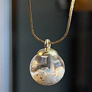 May include: A gold-colored necklace featuring a clear, spherical pendant. The pendant contains small, light-colored inclusions. The necklace chain is a thin, textured design. The pendant is suspended from a gold-colored bail.