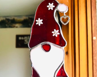 Op de afbeelding: Een gebrandschilderd glazen kabouterornament met een rode hoed versierd met sneeuwvlokaccenten en een witte baard. De kabouter heeft een zilveren hartvormige bedel die aan zijn hoed hangt. Het ornament is ontworpen om op te hangen.