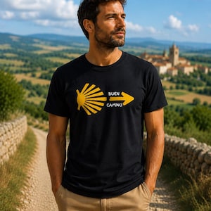 May include: A black short-sleeved t-shirt featuring a yellow graphic of a scallop shell and an arrow pointing right, with the text "BUEN CAMINO" printed in white letters between the shell and the arrow, centered on the chest of the garment.